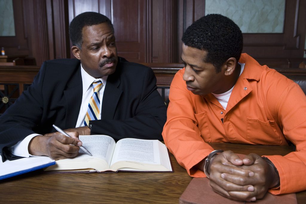  A criminal defense lawyer sits next to his client in an orange jumpsuit at a defense table in a courtroom.